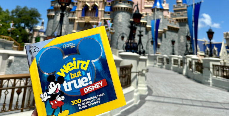 A copy of Weird But True! Disney is held up in front of Sleeping Beauty Castle at Magic Kingdom Park. The cover of the book features the title superimposed over a blue, three-circle Mickey. To the left of the title, a classic version of Mickey Mouse points to himself with a hand on his hip.