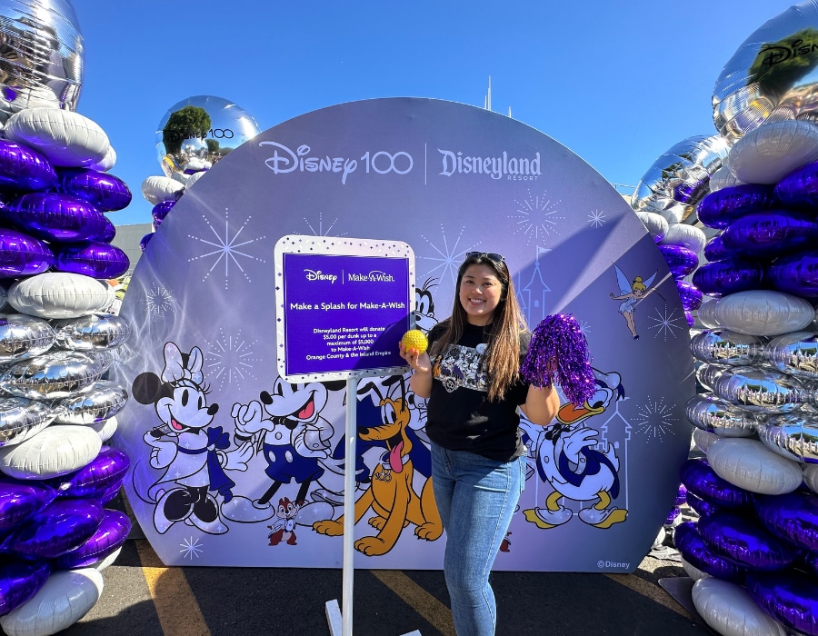 A cast member stands in front of a Disney100 photo backdrop and balloons, holding a plastic ball and cheerleading pom-pom