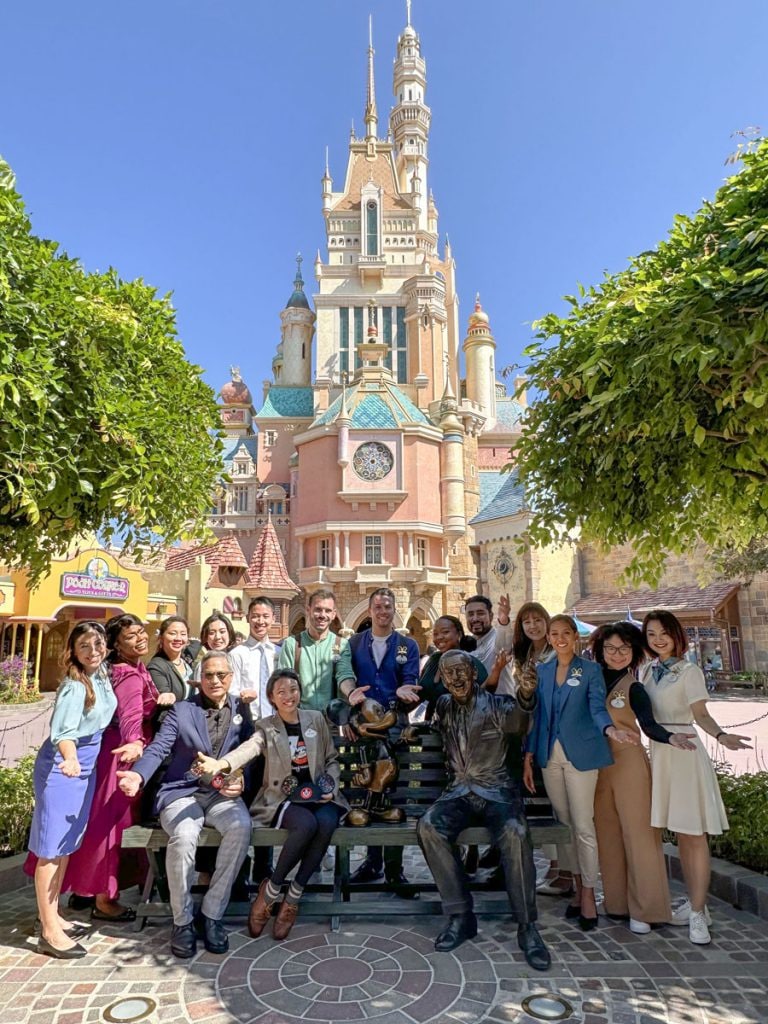 The Disney Ambassadors gather with Imagineers around the Dream Makers statue of Mickey Mouse and Walt Disney on a park bench, with the Castle of Magical Dreams behind them
