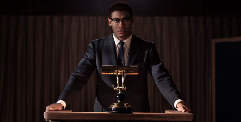 In Genius: MLK/X, Malcolm X, played by Aaron Pierre, stands at a podium.