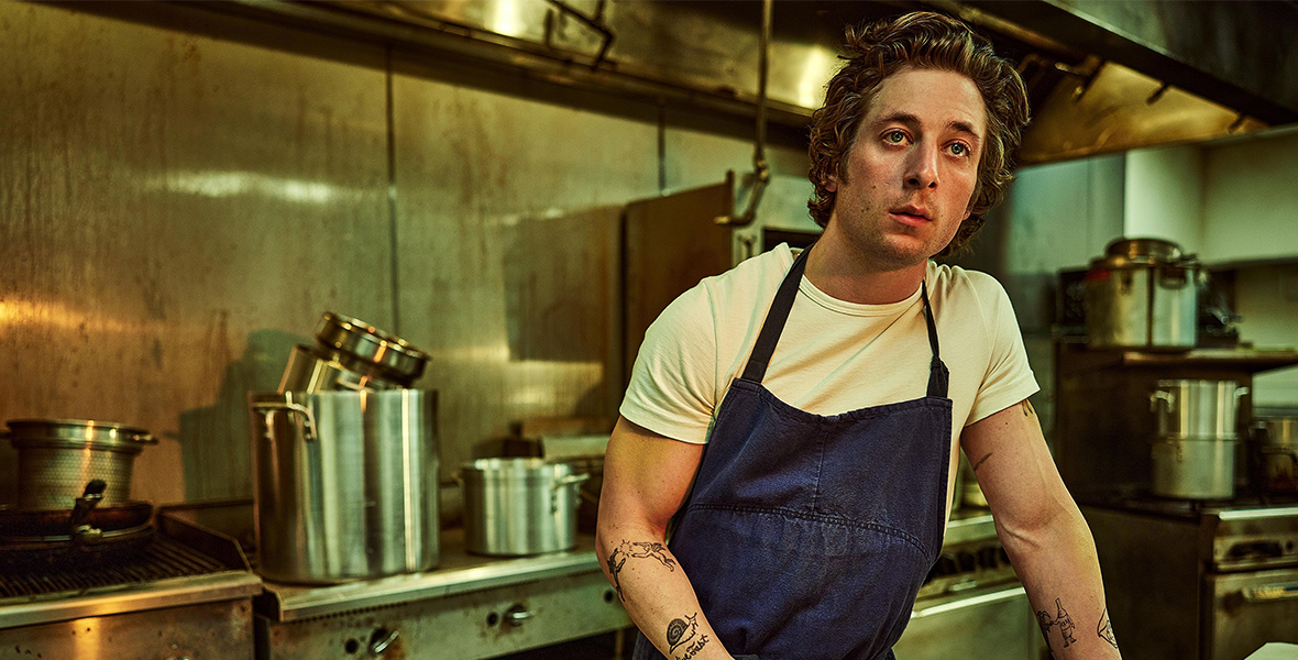 In a scene from the television show The Bear, Carmy, played by Jeremy White, stands in a restaurant kitchen. He wears a white T-shirt and a blue apron, holding a felt-tip pen in his right hand while appearing to write something on a pizza box. Courtesy of FX Networks.