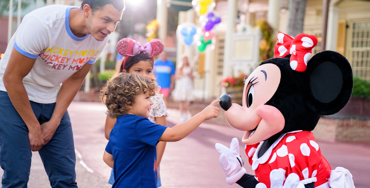 A father and two kids, a boy in a blue shirt and a girl wearing pink Minnie ears, are visiting with Minnie Mouse. Minnie is kneeling, and the boy is reaching out towards her nose.