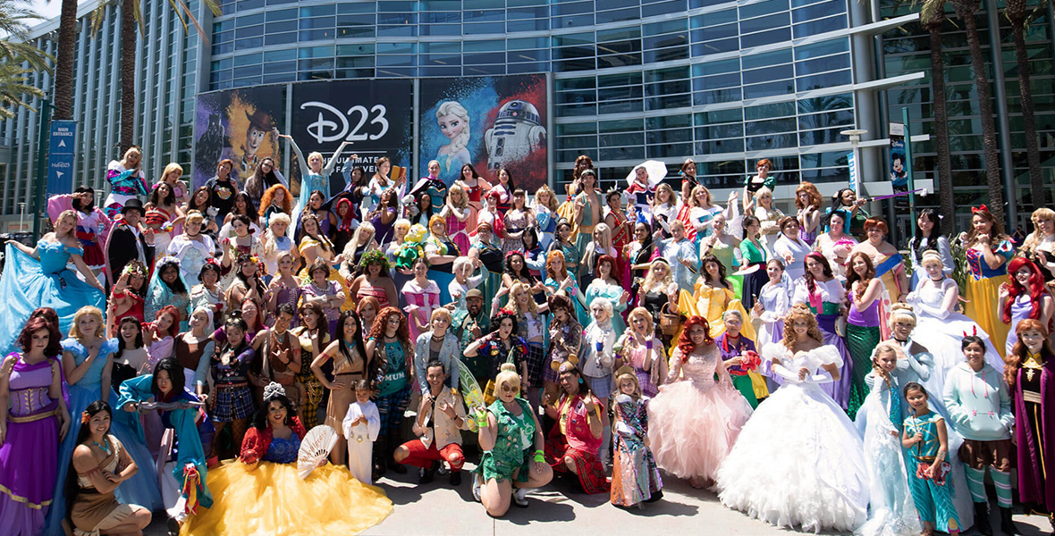 An image showing fans from a previous D23 Cosplay Meet-Up. Various fans of Disney Princesses franchises stand and kneel in front of the camera to showcase their cosplay creations in front of the Anaheim Convention Center. On the building is a billboard with the writing “D23 The Ultimate Disney Fan Event.”