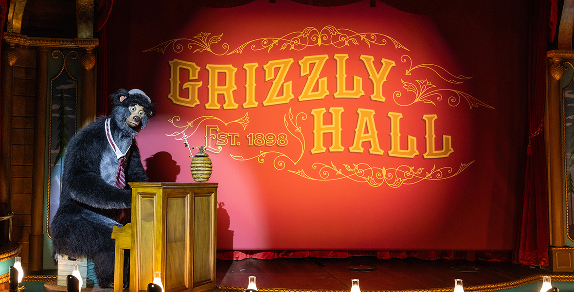 In an image from Walt Disney World’s Country Bear Musical Jamboree, on a vaudeville style stage is a tall grizzly bear seated at a small upright piano on the left. The bear is Gomer, and he wears a white collar with a red-and-blue-striped tie and a lavender hat. Atop the piano is a round beehive with two straws sticking out of the to. At the back of the stage is a red curtain with writing on it that says “Grizzly Hall, Established 1898.” Two spotlights illuminate Gomer and the curtain.