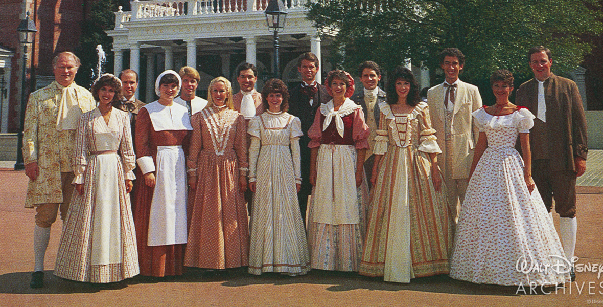 The Voices of Liberty a capella vocal group stands in two lines, shoulder to shoulder, in front of the American Adventure pavilion at EPCOT in the 1980s. In the front row are seven women, while eight men stand behind them. All are dressed in American Colonial costumes, with ankle-length dresses and a few aprons and caps on the women and knee-length socks worn below loose pants gathered below the knee on the men, who also sport ascots, vests, and jackets.