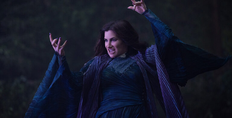 Agatha Harkness (Kathryn Hahn) from Marvel Television’s Agatha All Along wears a long-sleeved, pleated dark blue dress with dark purple fabric draped around her neck. She is pictured using her powers with slightly curled hands and purple-painted nails, speaking against a blurred, dark-toned background.