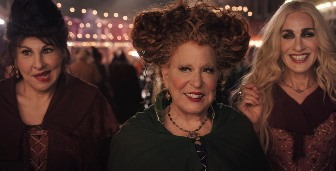 In a scene from Hocus Pocus 2 (from left to right), Mary Sanderson (Kathy Najimy), Winifred Sanderson (Disney Legend Bette Midler), and Sarah Sanderson (Sarah Jessica Parker) are standing, looking slightly to their right. Mary and Sarah are wearing burgundy cloaks, and Winifred wears a green cloak. In the background are outdoor string lights.