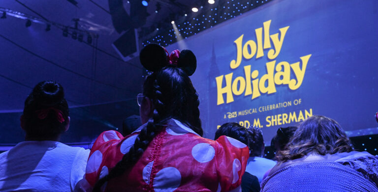 Seen from the perspective of the audience is part of the stage for the “Jolly Holiday: A D23 Musical Celebration of Richard M. Sherman” during D23: The Ultimate Disney Fan Event 2024. The logo for the show can be seen on the stage’s large screen backdrop, and in the foreground is an audience member wearing Minnie Mouse ears and a red polka-dot dress.