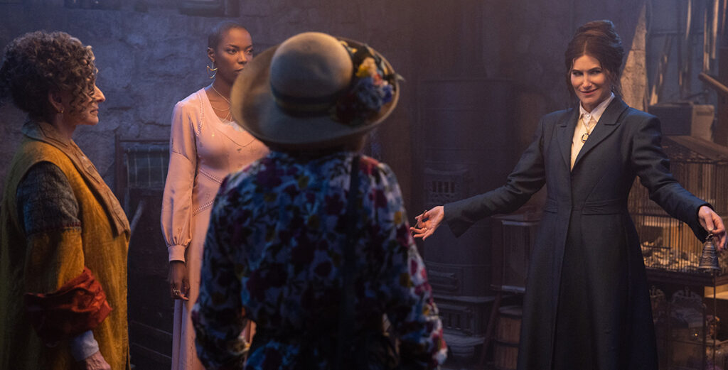 In a scene from Agatha All Along, Lilia Calderu (Patti LuPone), Jennifer Kale (Sasheer Zamata), Mrs. Hart/Sharon Davis (Debra Jo Rupp), and Agatha Harkness (Kathryn Hahn) are gathered in a stone-walled basement. Lilia wears a mustard and blue long-sleeve dress, Jennifer dons a long-sleeve baby pink dress, Mrs. Hart/Sharon Davis sports a floral long-sleeve dress, and a straw hat adorned with flowers, and Agatha Harkness is dressed in a navy blue, cinched long-sleeve coat over a white shirt.