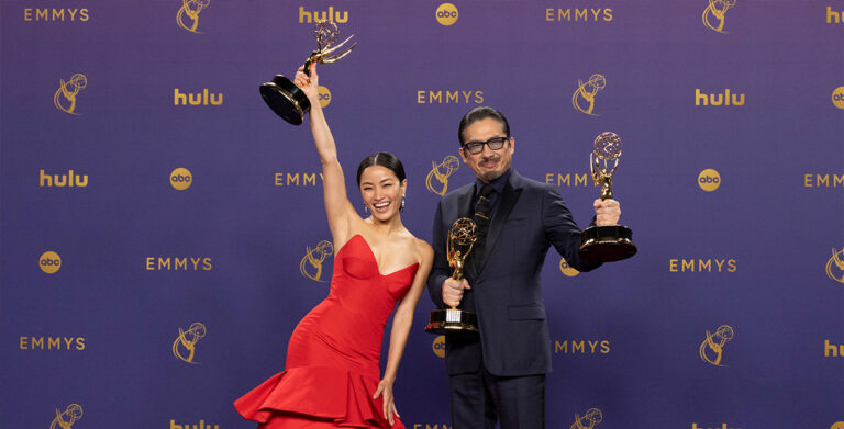 A celebratory image featuring two actors holding Emmy Awards at the Emmy Awards ceremony. On the left is Anna Sawai in a striking red gown joyfully raises her Emmy high. On the right is Hiroyuki Sanada in a classic dark suit proudly holds his Emmy.