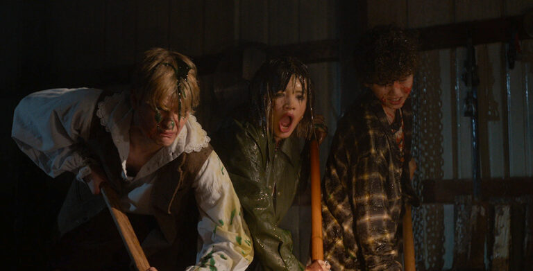 : In a first-look image from Carved (2024), (L-R) Corey Fogelmanis, Peyton Elizabeth Lee, and Wyatt Lindner, covered in colorful slime, hold shovels while digging. Their faces show shock and disgust. The dimly lit interior features wooden plank walls and metallic handles in the background.