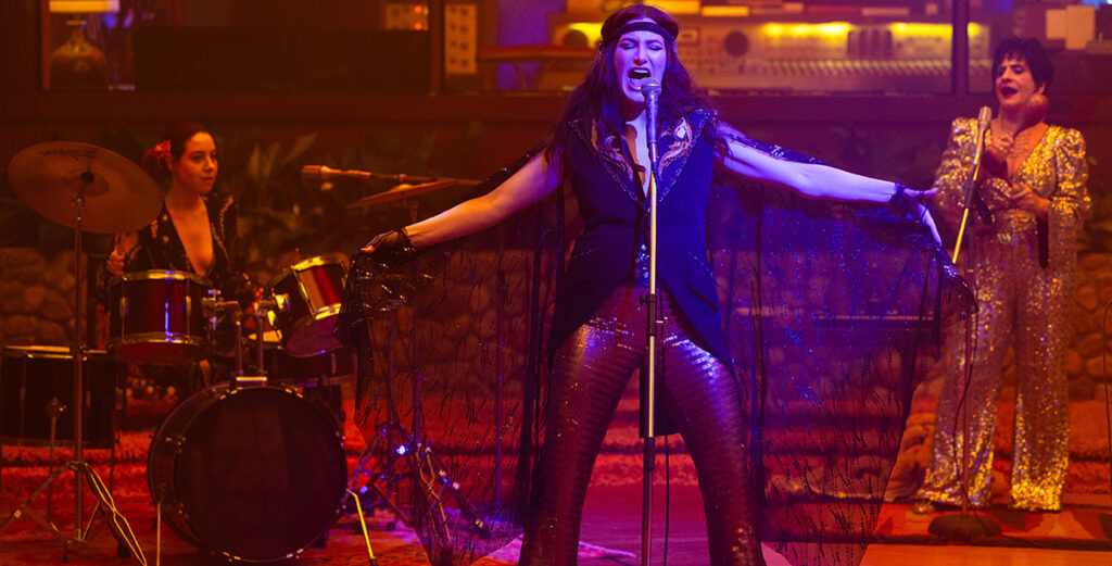 In a scene from Agatha All Along, we see from left to right Rio Vidal (played by Aubrey Plaza), Agatha Harkness (played by Kathryn Hahn), and Lilia Calderu (played by Patti LuPone) perform as a band. Rio plays the drums, wearing an animal-print, V-neck long-sleeve dress with a pink flower behind her right ear. Agatha sings, dressed in a headband, a V-neck black vest with gold details, pleated flare pants, and holding a black shawl. Lilia plays the maracas, wearing a sparkling gold jumpsuit. The frame is tinted with red and purple hues from the set’s lighting, and the background features a recording studio booth and a stone wall.