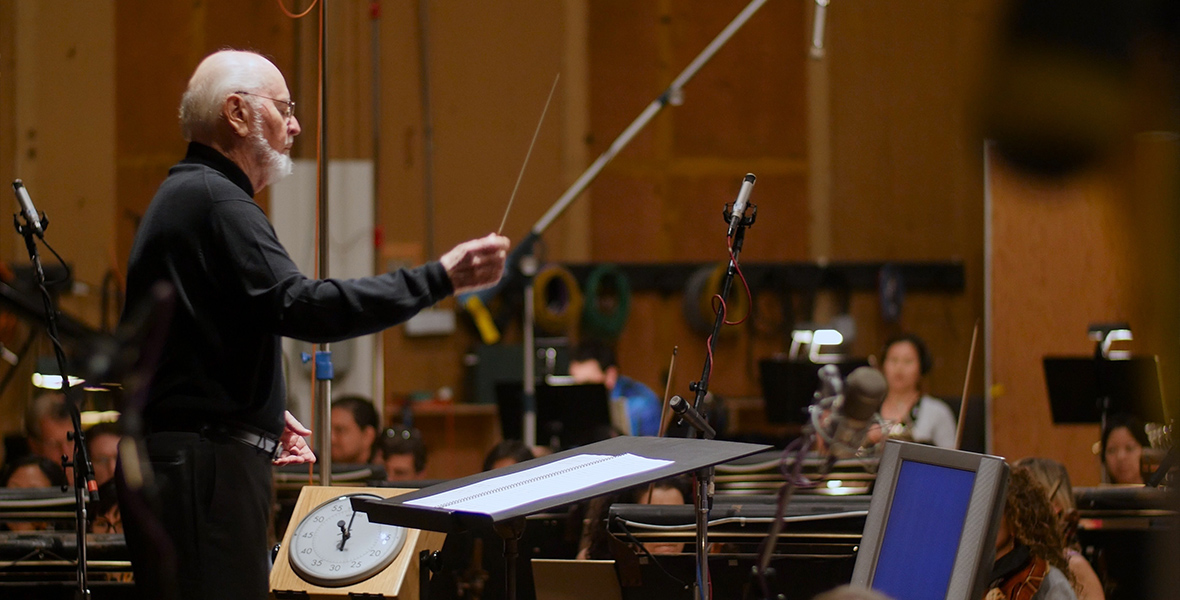 In an image from the Music by John Williams documentary, John Williams is conducting an orchestra. He wears a black mock turtleneck sweater and black pants, holding a baton as he looks downward at a large music stand elevated on a black podium. The blurred background features the wooden interior of the room, with microphones and orchestra members playing instruments, including violins.