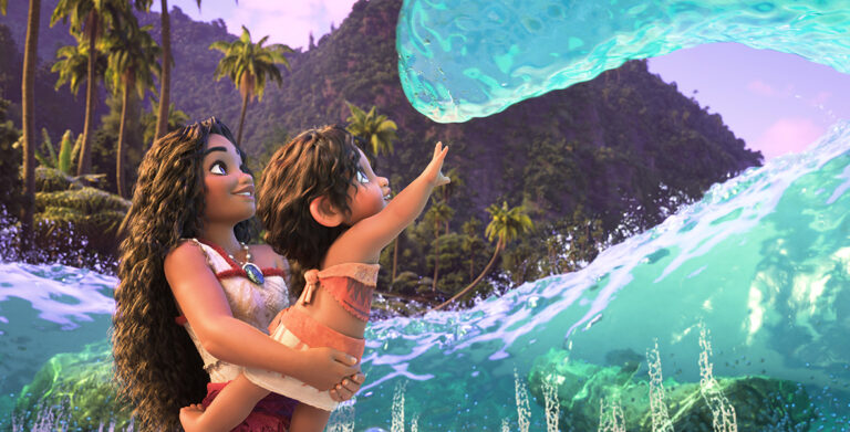 In an image from Walt Disney Animation Studios’ Moana 2, Moana (voiced by Auli‘i Cravalho) is holding her little sister Simea (voiced by Khaleesi Lambert-Tsuda) up to touch a wave; they are standing on a Motunui beach, surrounded by the water, which is acting friendly to the pair as it has to Moana in the past. There are lush green mountains and palm trees seen in the background.