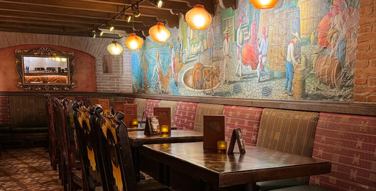 An image of the La Cava del Tequila restaurant interior. Rustic and cozy with circular lamps hang down from the ceiling and wooden chairs are pushed into wooden candlelit tables. The Mexican tequila bar features a unique handmade mural over a brick wall, depicting the process of the agave spirits distillation—from the harvesting and the cooking, to the fermentation, distillation, and aging in the big brown barrels.