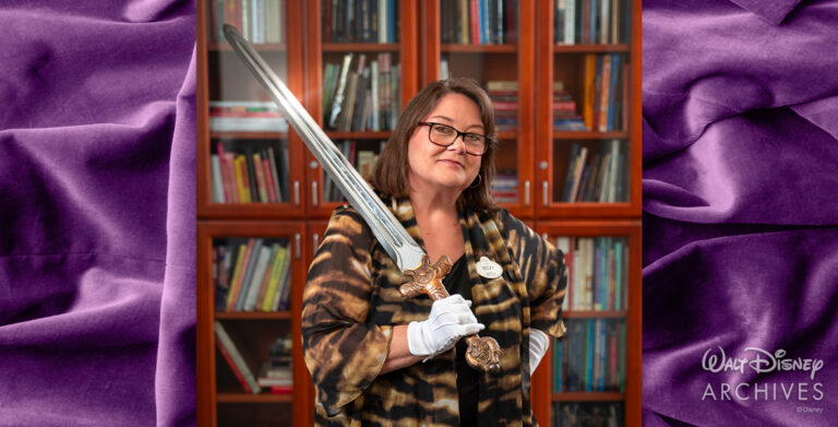 Walt Disney Archives director Becky Cline, wearing a patterned shawl and white gloves, poses confidently while holding the “Excalibur,” a decorative prop sword from the 2004 film King Arthur, with shelves filled with books in the background.